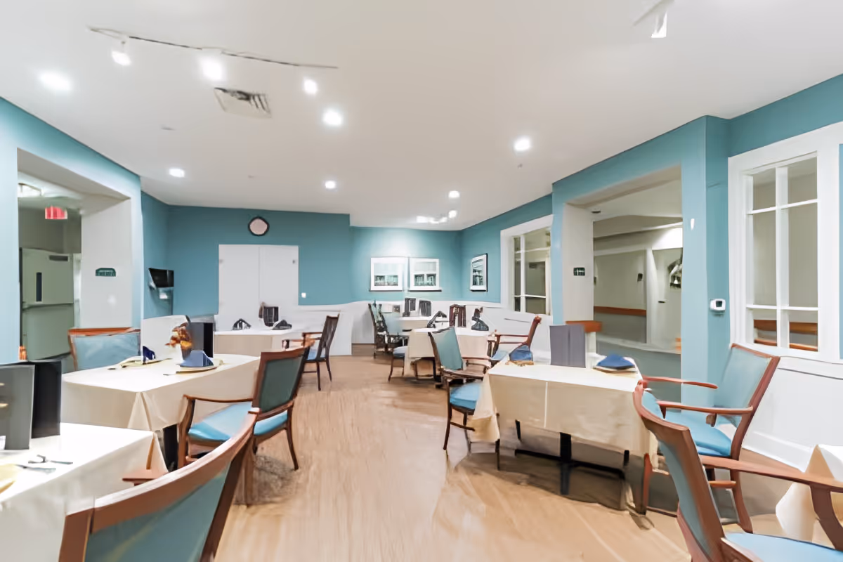 Interior view of a dining room in a senior living facility with multiple tables covered with beige tablecloths and set with napkins and utensils. The walls are painted teal, and there are wooden chairs with teal cushions around the tables. The room is well-lit with ceiling lights, and there are windows and doorways visible along the walls.
