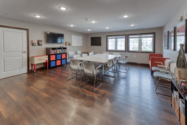 Well-lit common activity/dining room with tables and chairs, a wall-mounted TV, bookshelves, and windows.