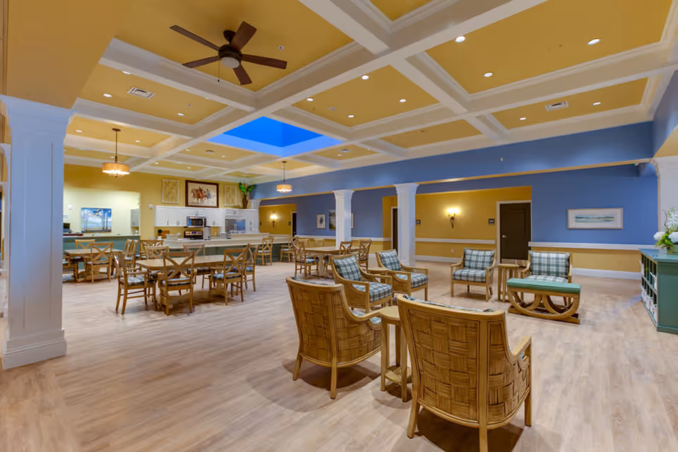Spacious senior living common area with dining tables, lounge chairs, and a kitchen under a coffered ceiling with a skylight.