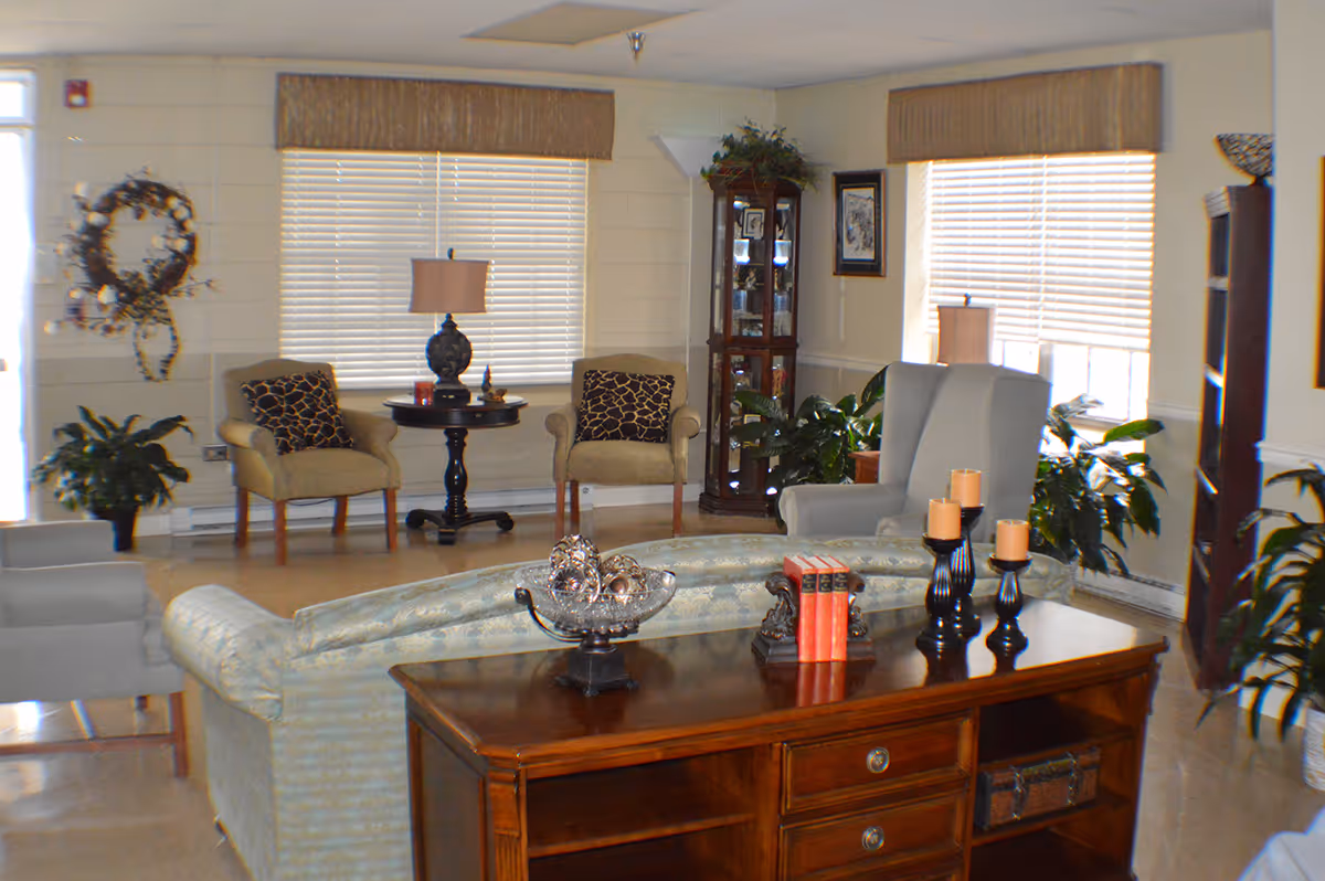 A cozy living room area with a light blue patterned sofa, a wooden console table with decorative items including candles and books, two beige armchairs with patterned cushions, a round side table with a lamp, a tall glass display cabinet, several potted plants, and windows with blinds and valances letting in natural light.