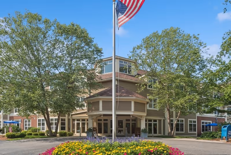 Exterior view of The Gables at Winchester senior living facility with a circular driveway, landscaped flower bed in the foreground, two large trees, and an American flag on a flagpole in front of the building under a clear blue sky.