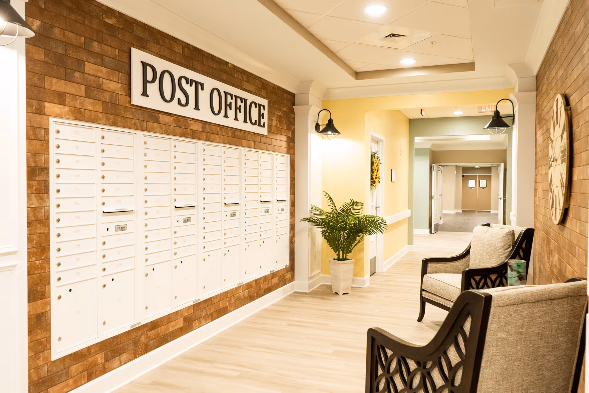 Interior hallway of a senior living facility with a wall of white mailboxes labeled POST OFFICE. The hallway has light wood flooring, yellow and green painted walls, two cushioned chairs with dark wooden frames, a large wall clock, a potted plant, and overhead lighting fixtures.