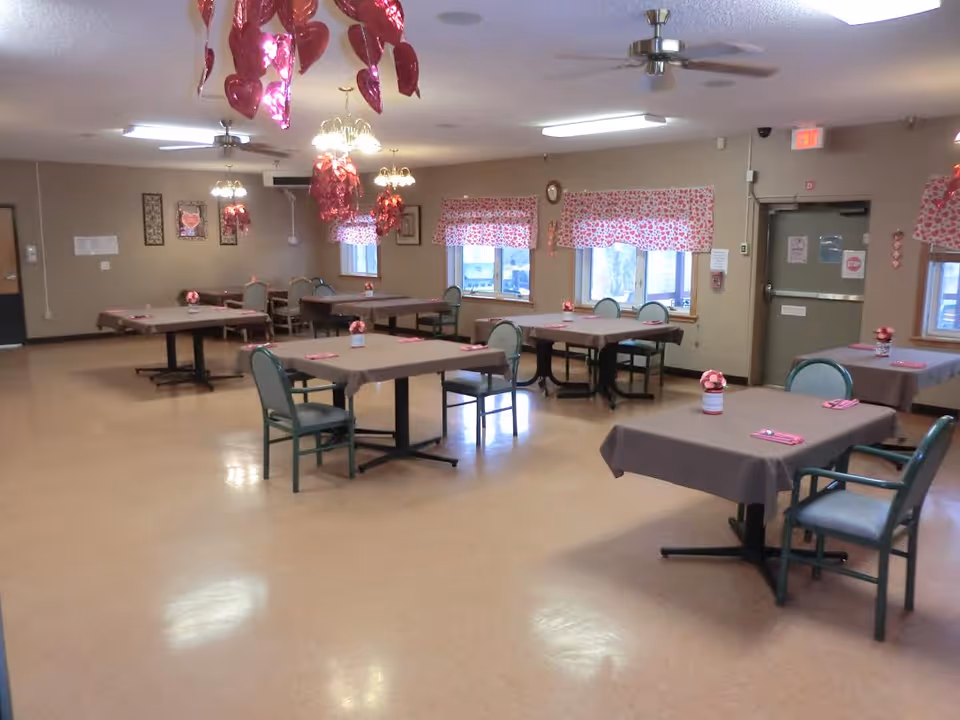 A dining room in a senior living facility with several tables covered in brown tablecloths, each set with pink napkins and small flower centerpieces. The room has beige walls, windows with red and white patterned valances, ceiling fans, and hanging red heart-shaped decorations.