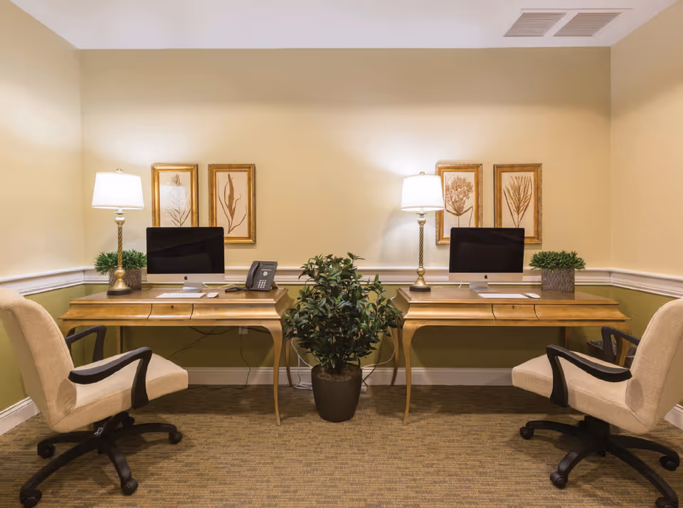 Two wooden desks with computers, lamps, and swivel chairs facing each other in a warmly lit interior workspace with a potted plant between them.
