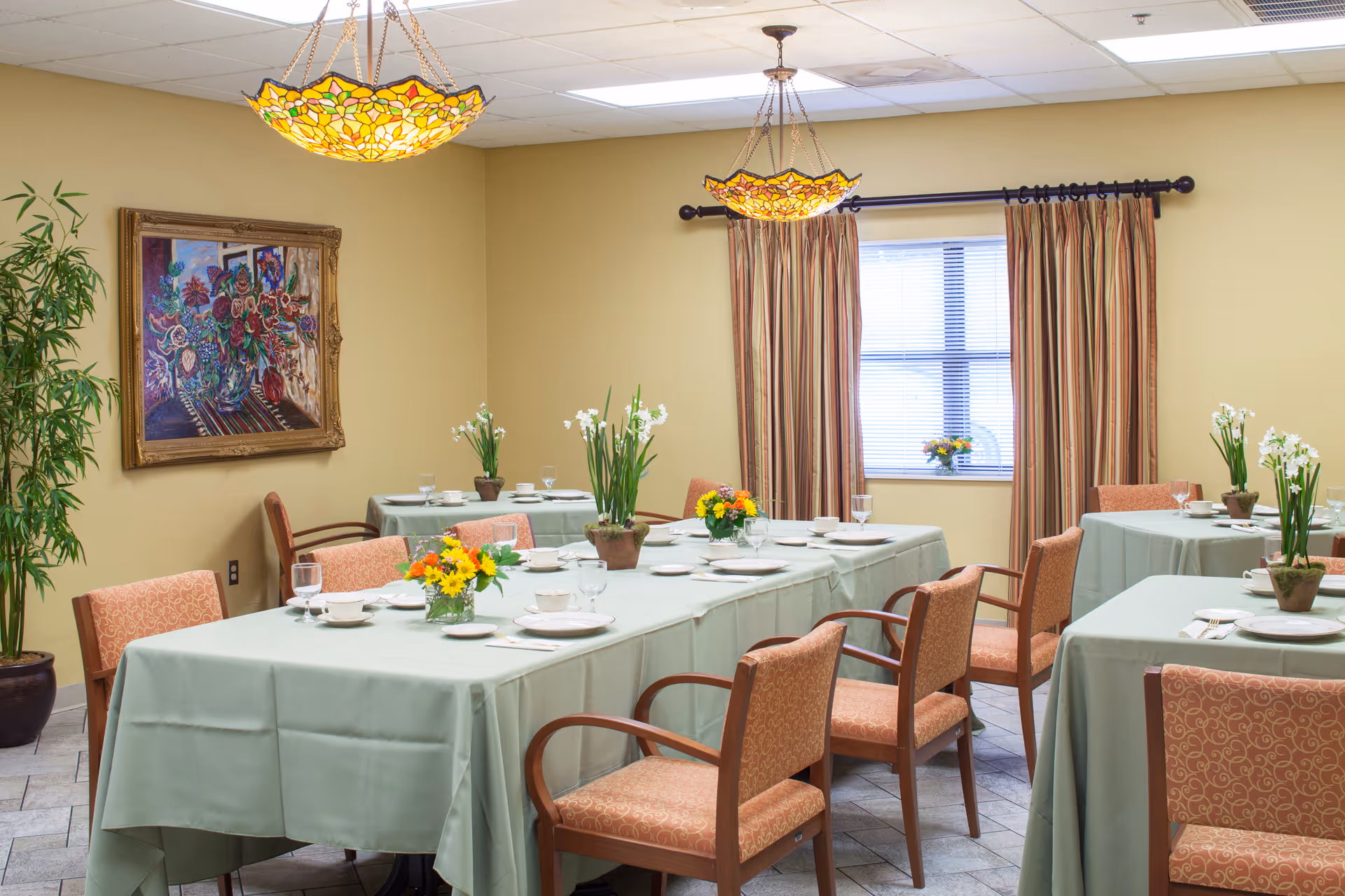 Dining room with multiple tables draped in light green tablecloths, floral centerpieces, upholstered chairs, and stained-glass pendant lights.