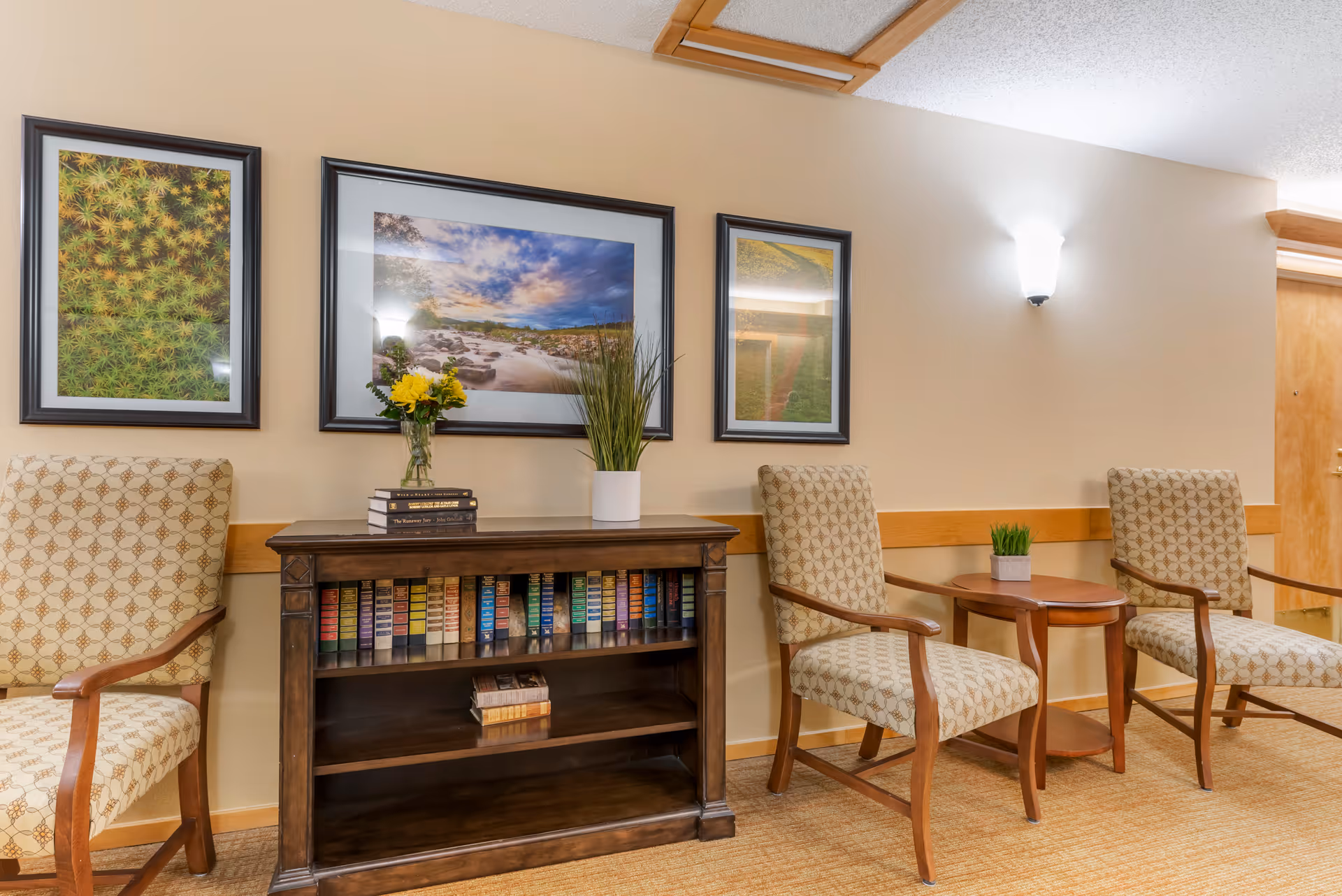 Cozy seating area with patterned armchairs, a wooden bookshelf filled with books and decorative plants, and framed landscape artwork on the wall.