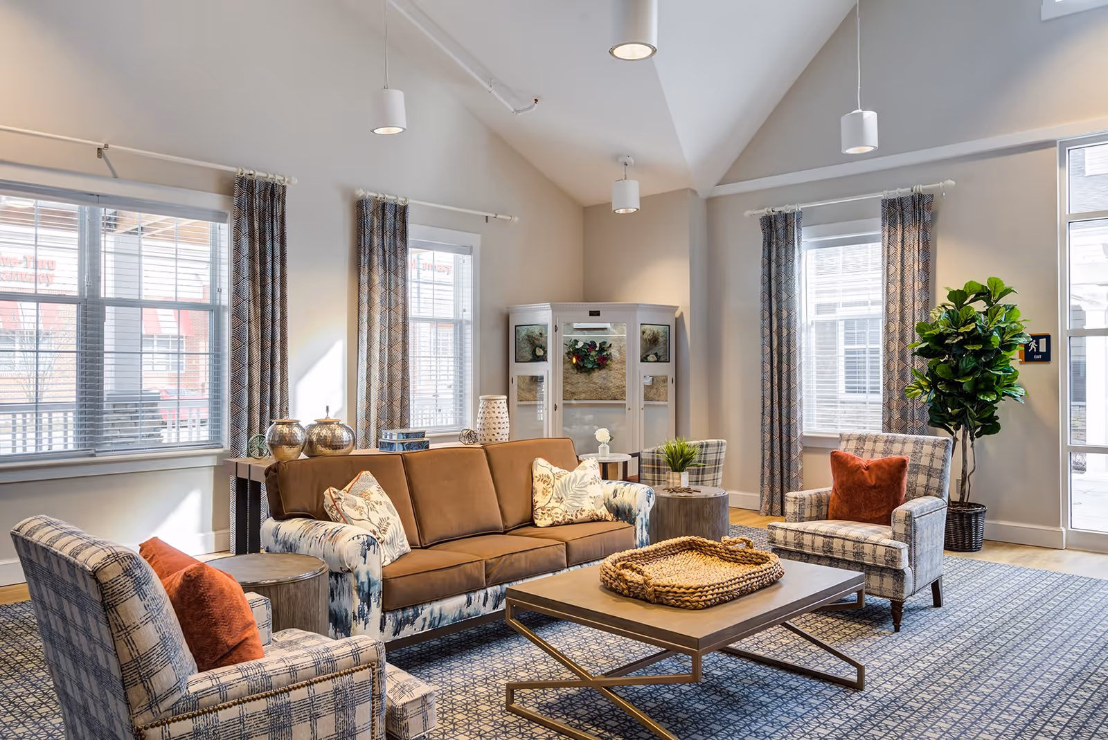 A bright and cozy living room area with a brown sofa, two patterned armchairs with orange cushions, a wooden coffee table with a woven tray, and a patterned rug. The room has large windows with patterned curtains, a tall green plant in a basket, and soft overhead lighting.