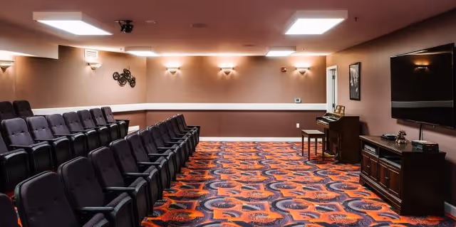 A small theater or media room with two rows of black cushioned seats facing a large flat-screen TV mounted on the wall. The room has patterned carpet in shades of orange, purple, and red, beige walls with wall sconces providing soft lighting, and a piano with a bench near the TV.