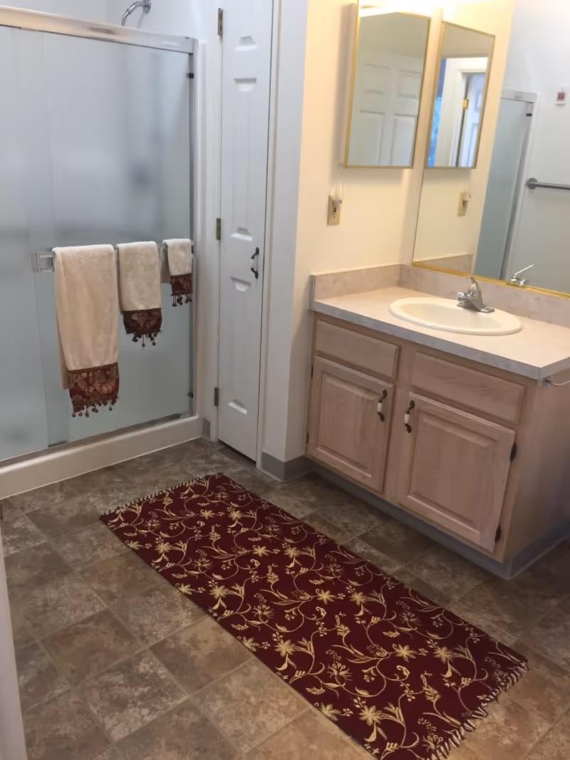 Bathroom with a frosted glass shower, vanity with sink and mirror, and a red patterned runner on the tile floor.