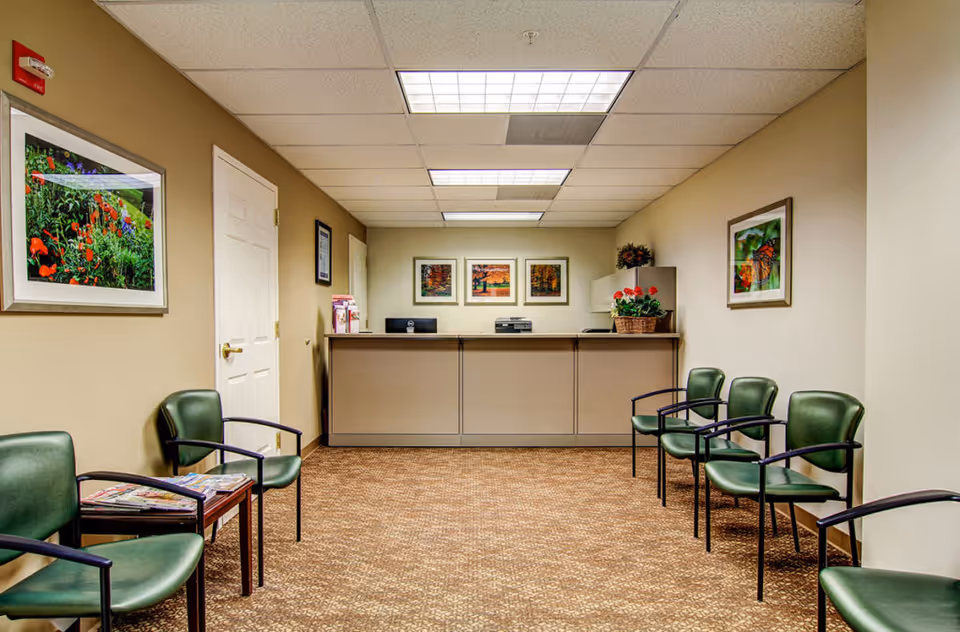 Reception area with a front desk at the end of the room, flanked by green chairs arranged along both walls. The walls are decorated with framed pictures of flowers and nature scenes. The floor is carpeted, and the ceiling has fluorescent lighting panels.