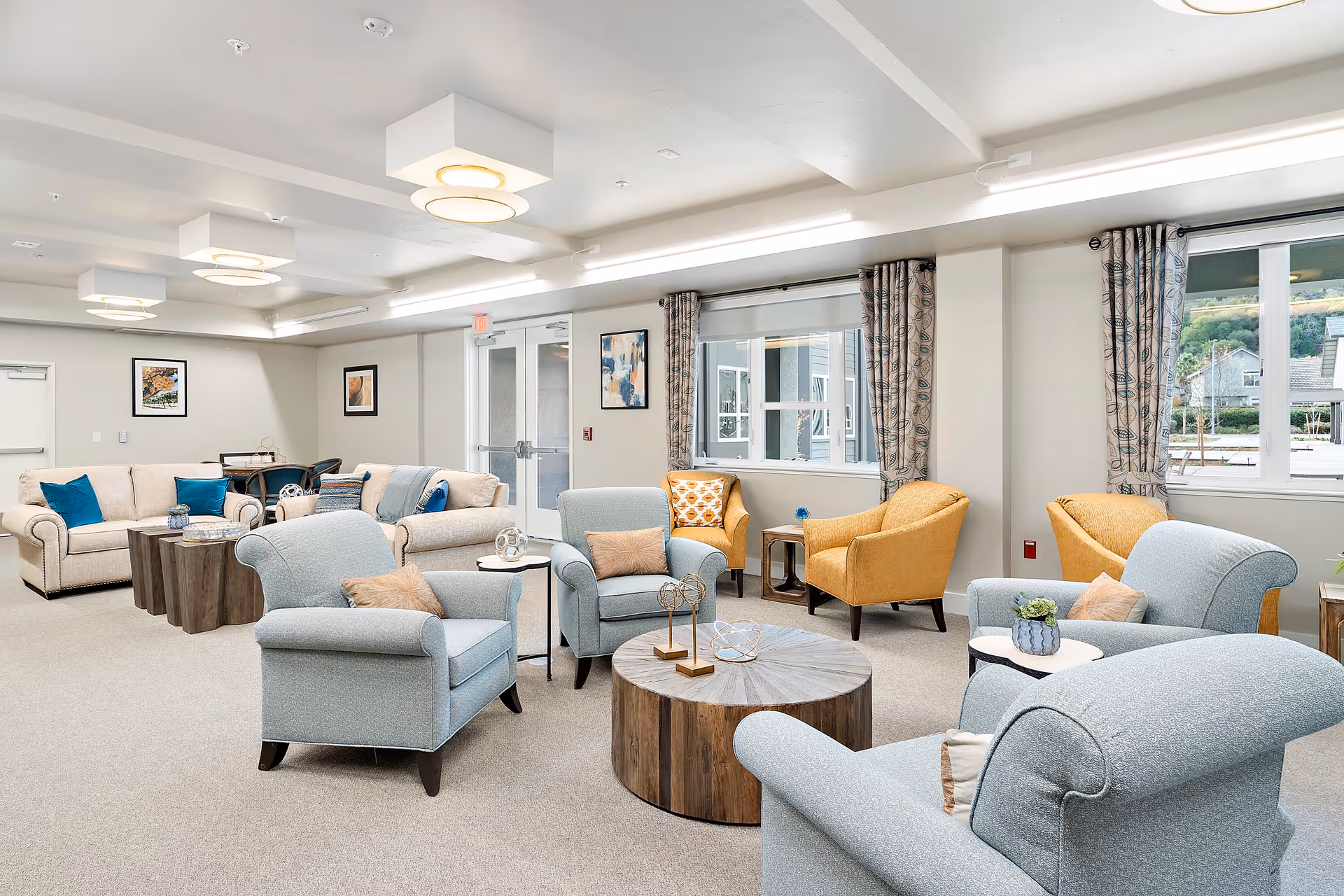 A bright and spacious living room area with light blue armchairs, beige sofas with blue cushions, and yellow armchairs arranged around wooden coffee tables. The room has large windows with patterned curtains, modern ceiling lights, and framed artwork on the walls.