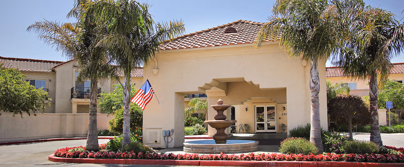 The entrance of Vista Harden Ranch featuring a decorative fountain, palm trees, and an American flag, surrounded by colorful flowers.