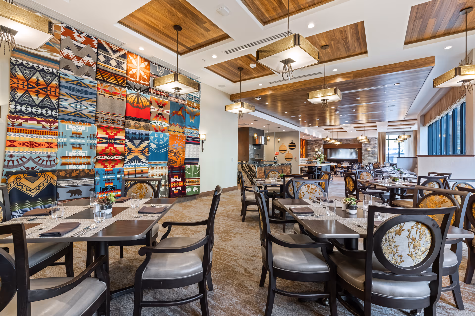 A spacious and well-lit dining room in Aegis Living Mercer Island featuring multiple dark wood tables and chairs with patterned upholstery. The room has a decorative wall with colorful, geometric-patterned textile art. The ceiling has wooden paneling with modern rectangular pendant lights hanging down. Large windows on one side allow natural light to fill the space, and the tables are set with placemats, napkins, glasses, and small potted plants.