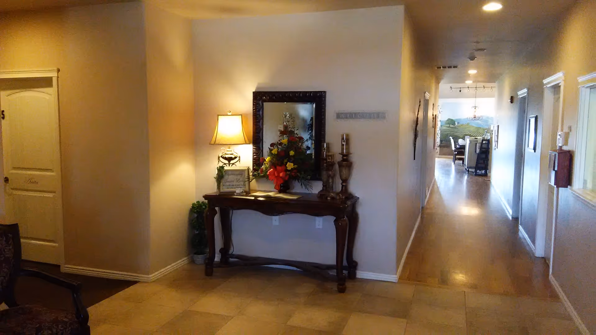 Interior hallway of an assisted living facility with a wooden console table against a wall. The table holds a lamp, a flower arrangement, candles, and a framed sign. A large mirror hangs above the table. The hallway extends to a dining area with a mural of a vineyard on the far wall. Doors and windows line the hallway walls.