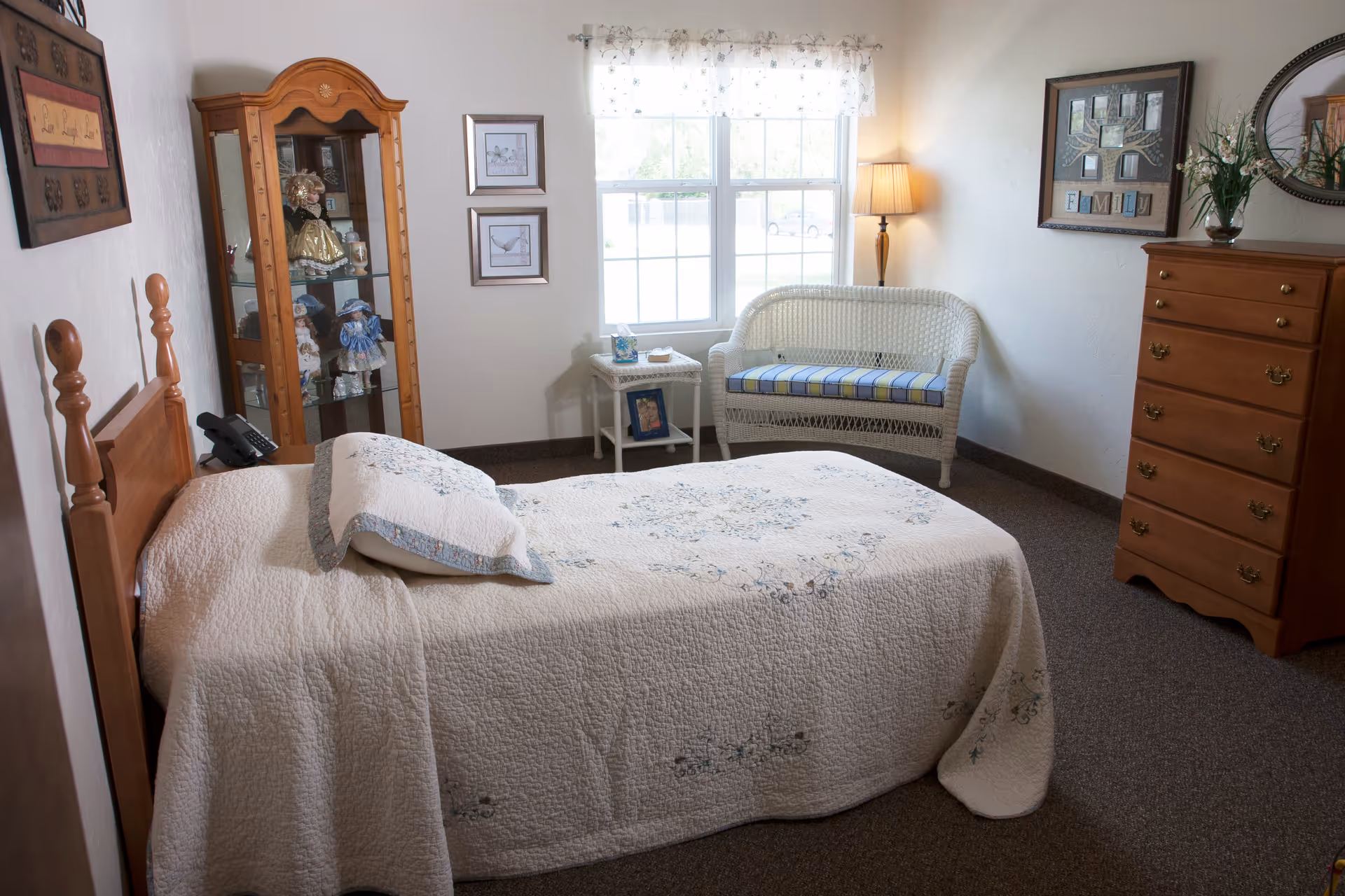 Sunlit bedroom with a single bed covered by a quilt, a wicker bench under the window, a wooden dresser, and a glass-front curio cabinet.