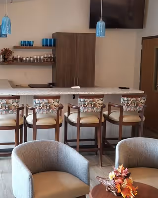 Communal lounge area with a bar counter and four patterned barstools, pendant lights, a wall-mounted TV, and nearby armchairs around a small table with fall floral decor.