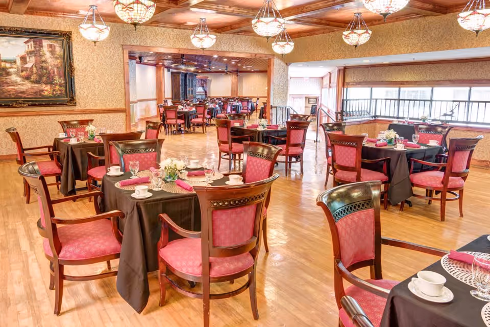 A spacious dining room with multiple round tables covered in black tablecloths, each set with white cups, glasses, and pink napkins. The chairs have wooden frames with pink upholstered seats and backs. The room features wooden flooring, patterned wallpaper, decorative ceiling lights, and a large painting on one wall.