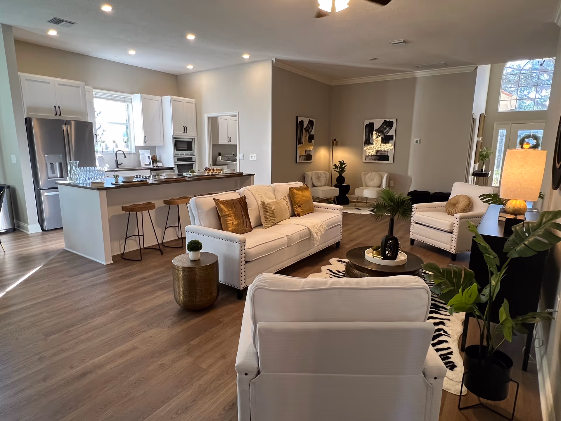 A bright and spacious open-concept living room and kitchen area in a senior living facility. The living room features white upholstered sofas and armchairs with gold and beige accent pillows, a round coffee table, and decorative plants. The kitchen has white cabinets, a stainless steel refrigerator, a kitchen island with three wooden bar stools, and various kitchen appliances. The room has wood flooring and recessed lighting.