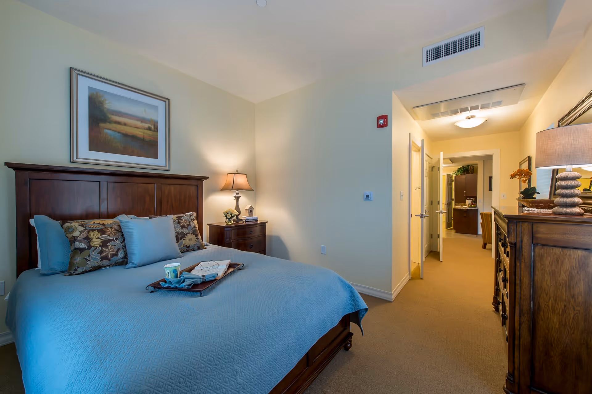A cozy bedroom with a large wooden bed featuring a blue bedspread and decorative pillows. A tray with a cup and a book is placed on the bed. There is a wooden nightstand with a lamp and a small plant beside the bed. On the wall above the bed hangs a framed landscape painting. The room has beige walls and carpeted floor. A hallway leads to another room with visible kitchen cabinets and dining chairs.