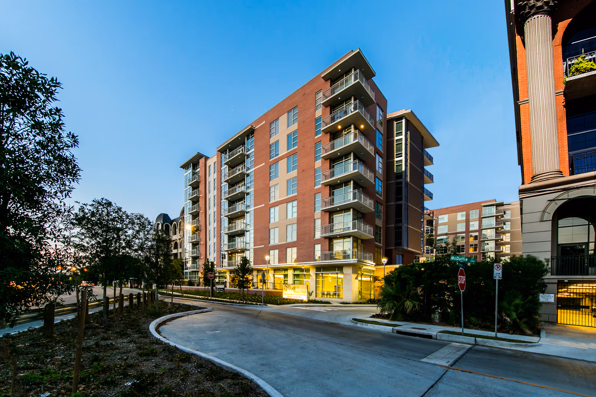 Exterior view of a modern multi-story residential building with balconies, surrounded by trees and a curved driveway, taken during early evening with clear sky.