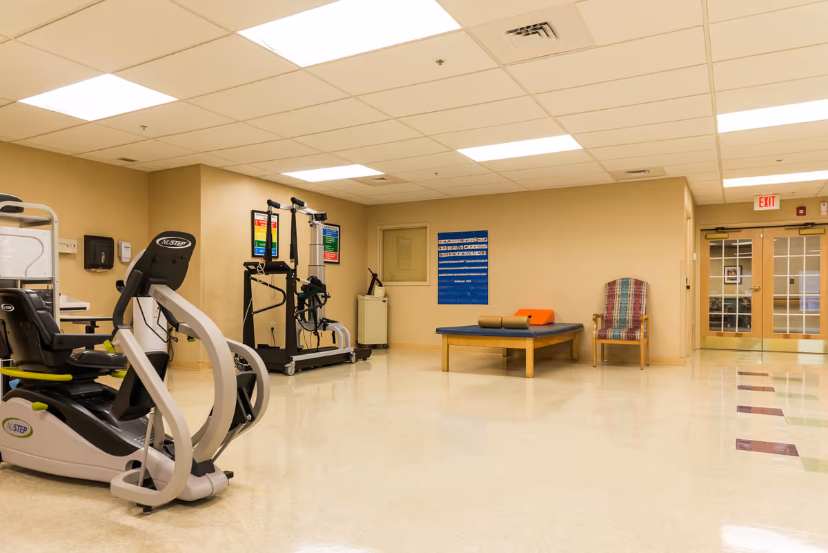 Well-lit therapy/exercise room with cardio machines, a treatment table, a chair, and double doors in the background.