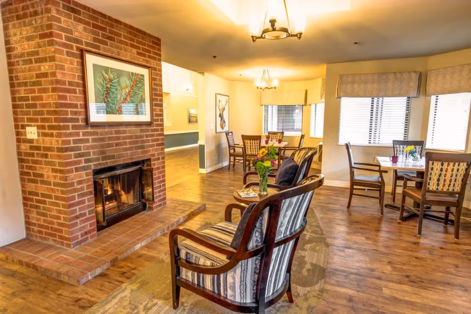 A cozy senior living common area with a brick fireplace on the left, two cushioned armchairs with wooden frames facing the fireplace, and small tables with flower vases. In the background, there are dining tables with chairs near windows with blinds and valances, and warm overhead lighting.
