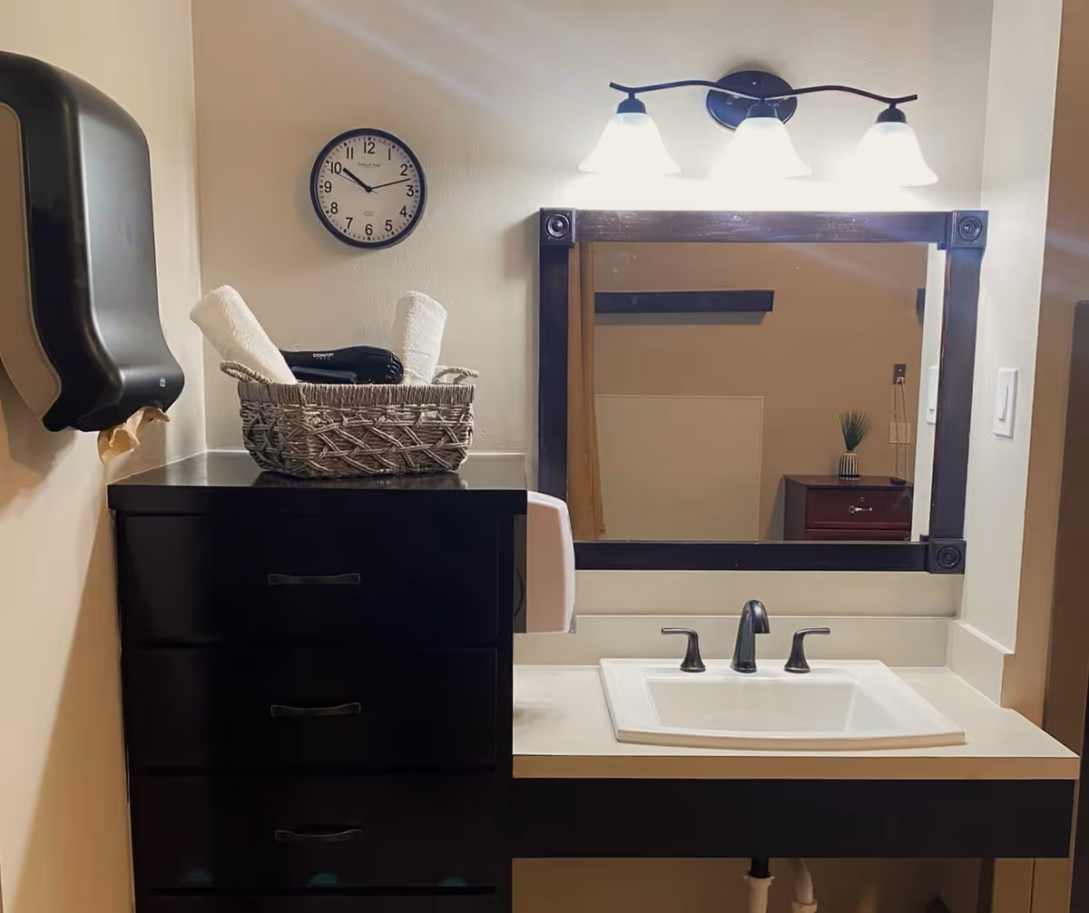 A bathroom vanity area with a white sink and black faucet, a large rectangular mirror with a dark wooden frame, and three light fixtures above the mirror. To the left, there is a black chest of drawers with a wicker basket on top containing rolled white towels and a black hairdryer. A wall clock shows the time as 9:10. A black paper towel dispenser is mounted on the wall to the left.