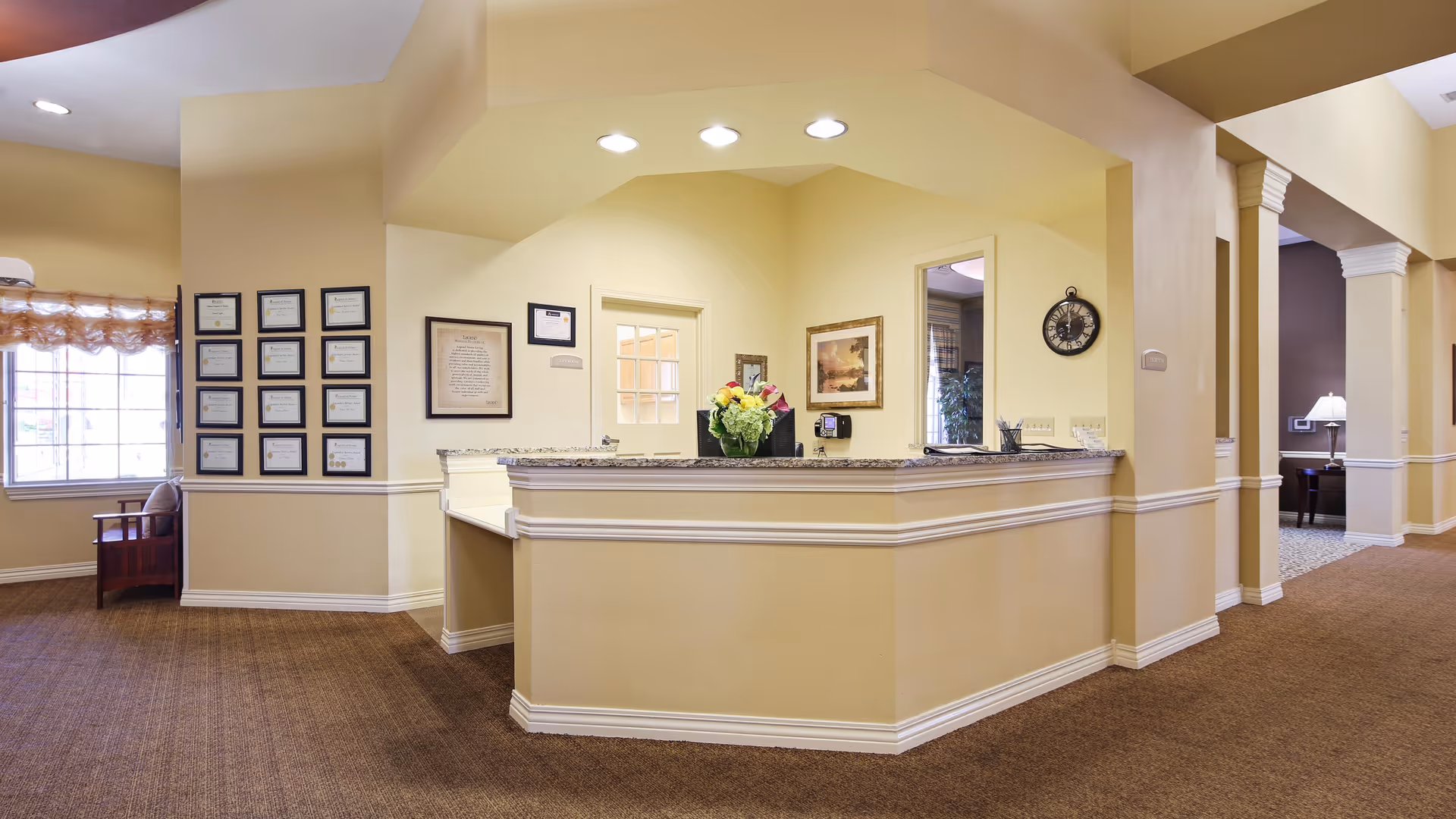 Reception area in a senior living facility with a beige and cream color scheme, a granite countertop desk with a flower arrangement, framed certificates and artwork on the walls, a clock, and a window with a chair nearby.