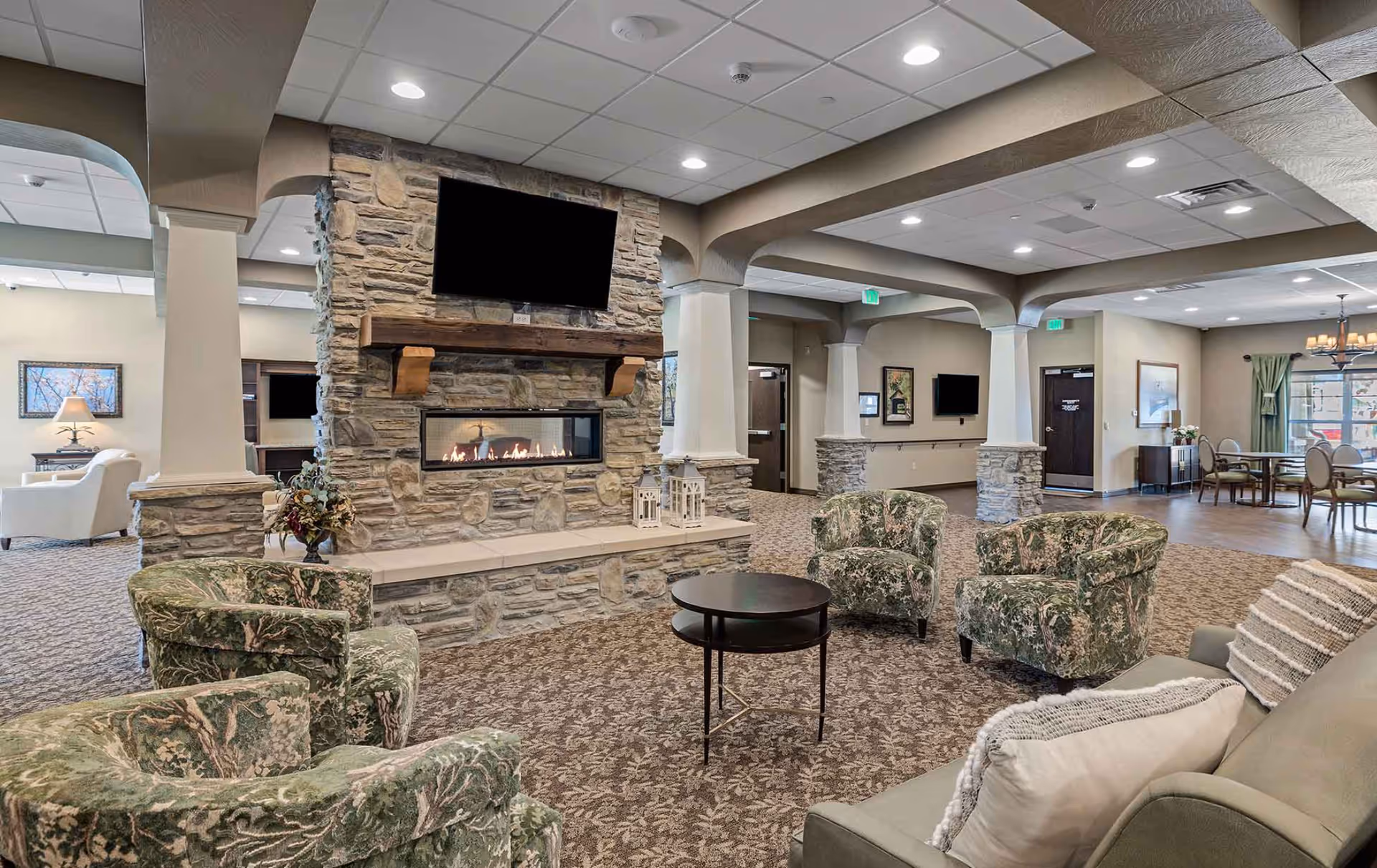 A spacious senior living facility common area with a stone fireplace and a mounted flat-screen TV above it. The room features patterned carpet, several upholstered armchairs with floral designs, a round black coffee table, and a sofa with pillows. In the background, there is a dining area with tables and chairs, and the space is well-lit with recessed ceiling lights.