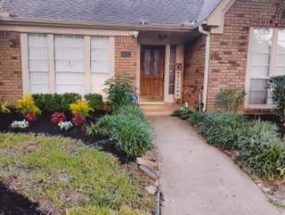 Front entrance of a brick house with a wooden door and a welcome sign next to it. The walkway leading to the door is bordered by green plants and shrubs, with a small garden area on the left side.
