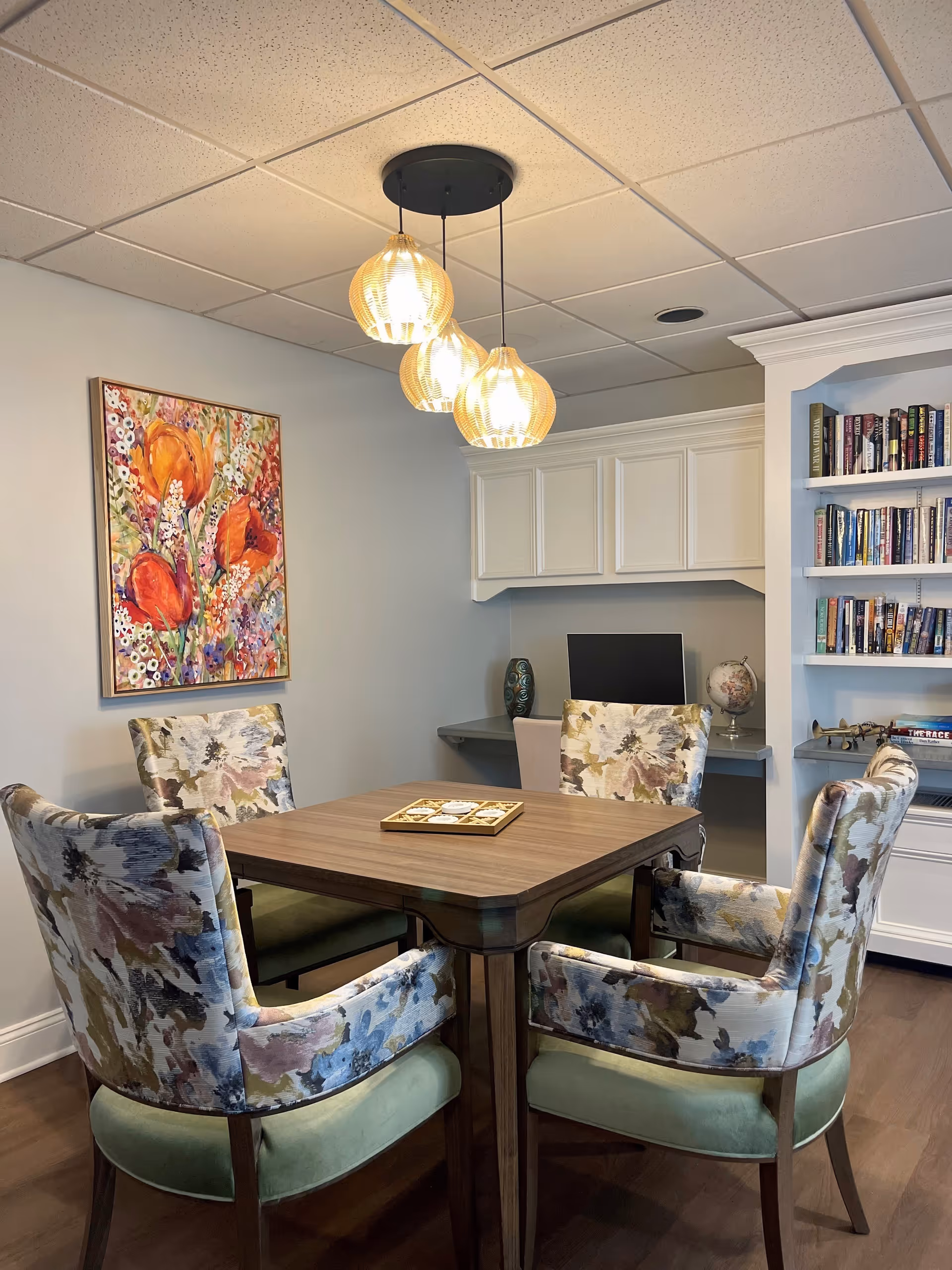 A cozy interior room with a square wooden table surrounded by four floral upholstered chairs with green cushions. Above the table hang three pendant lights with warm glowing bulbs. On the wall to the left is a colorful floral painting. In the background, there is a built-in desk with a computer monitor, a decorative vase, and a globe. To the right, a built-in bookshelf filled with books is visible.