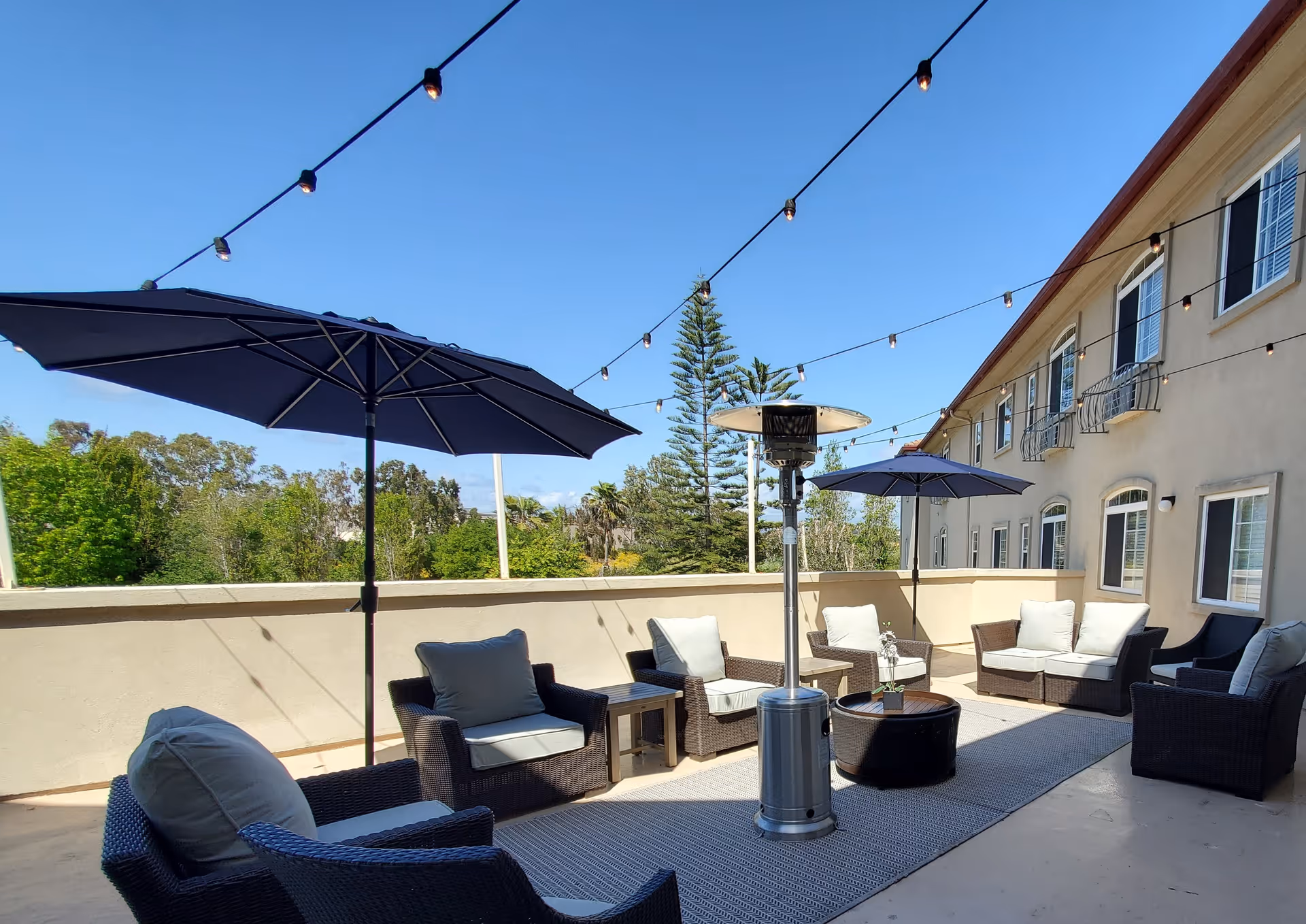 Outdoor patio area with cushioned wicker chairs and sofas arranged around a central heater and small tables. Two large navy blue umbrellas provide shade, and string lights are hung overhead. The patio is adjacent to a beige building with multiple windows and overlooks green trees under a clear blue sky.