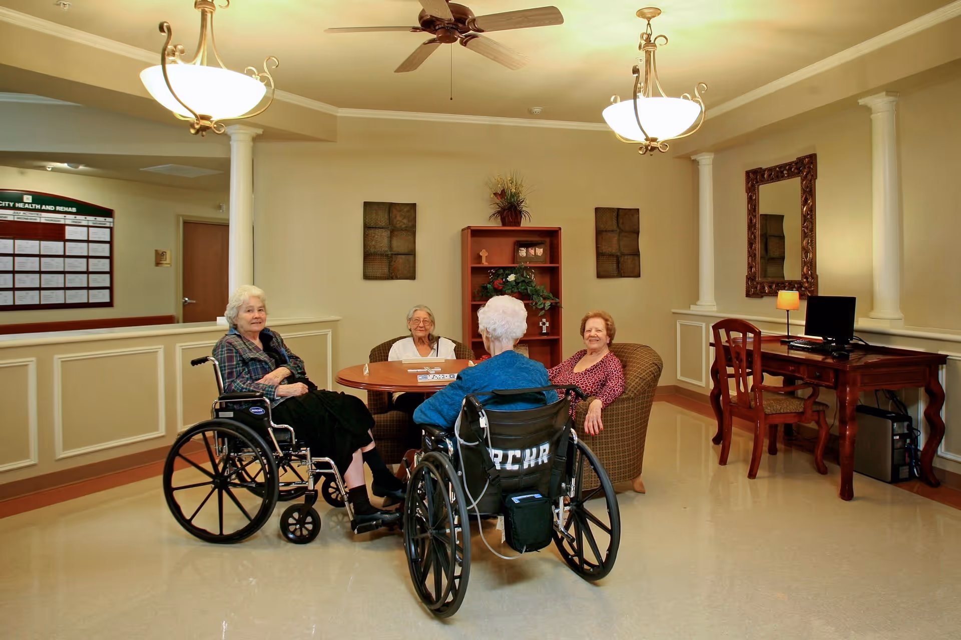 Four elderly women sitting around a round wooden table in a well-lit room with beige walls and decorative columns. Two women are in wheelchairs, and two are seated in armchairs. There is a wooden desk with a computer and chair on the right side of the room, and decorative wall art and a mirror on the walls.