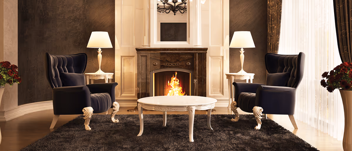 A cozy living room with two dark blue armchairs facing each other across a white round coffee table on a dark shaggy rug. Behind the table is a lit fireplace with a decorative mantel, flanked by two white side tables each holding a lamp with a white shade. There are large windows with sheer curtains on the right side, and flower arrangements in vases on both sides of the room.