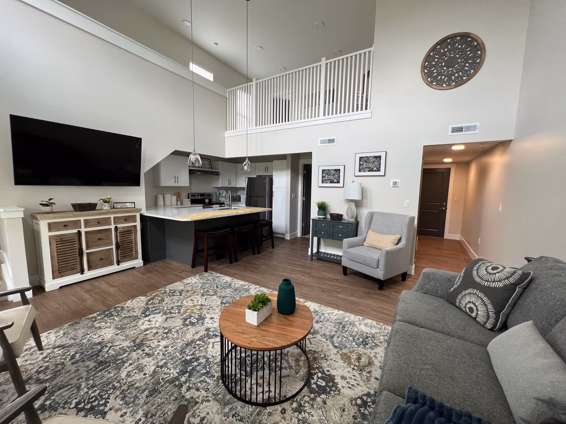 A modern living room and kitchen area with high ceilings and a loft railing above. The living room has a gray sofa with patterned pillows, a gray armchair, a round wooden coffee table with decorative items, and a patterned area rug. The kitchen features white cabinets, stainless steel appliances, a large island with a white countertop, and three bar stools. A flat-screen TV is mounted on the wall above a wooden cabinet. The walls are painted light gray, and there is a decorative round wall piece above the armchair.