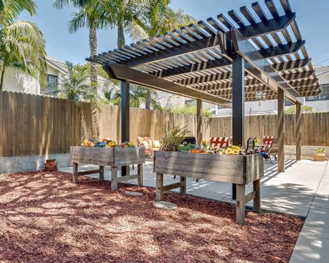 Outdoor patio area with a wooden pergola providing shade over seating with colorful cushions. Raised garden beds with flowers are in the foreground, surrounded by mulch and palm trees, with a wooden fence enclosing the space.