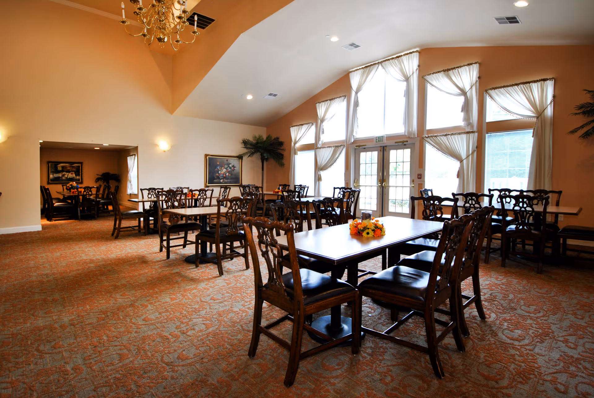 A spacious dining room with multiple wooden tables and chairs arranged neatly. The room features large windows with sheer curtains allowing natural light to fill the space. There are floral centerpieces on the tables and a chandelier hanging from the ceiling. The walls are painted in warm tones, and there is a painting and a potted plant visible in the background.