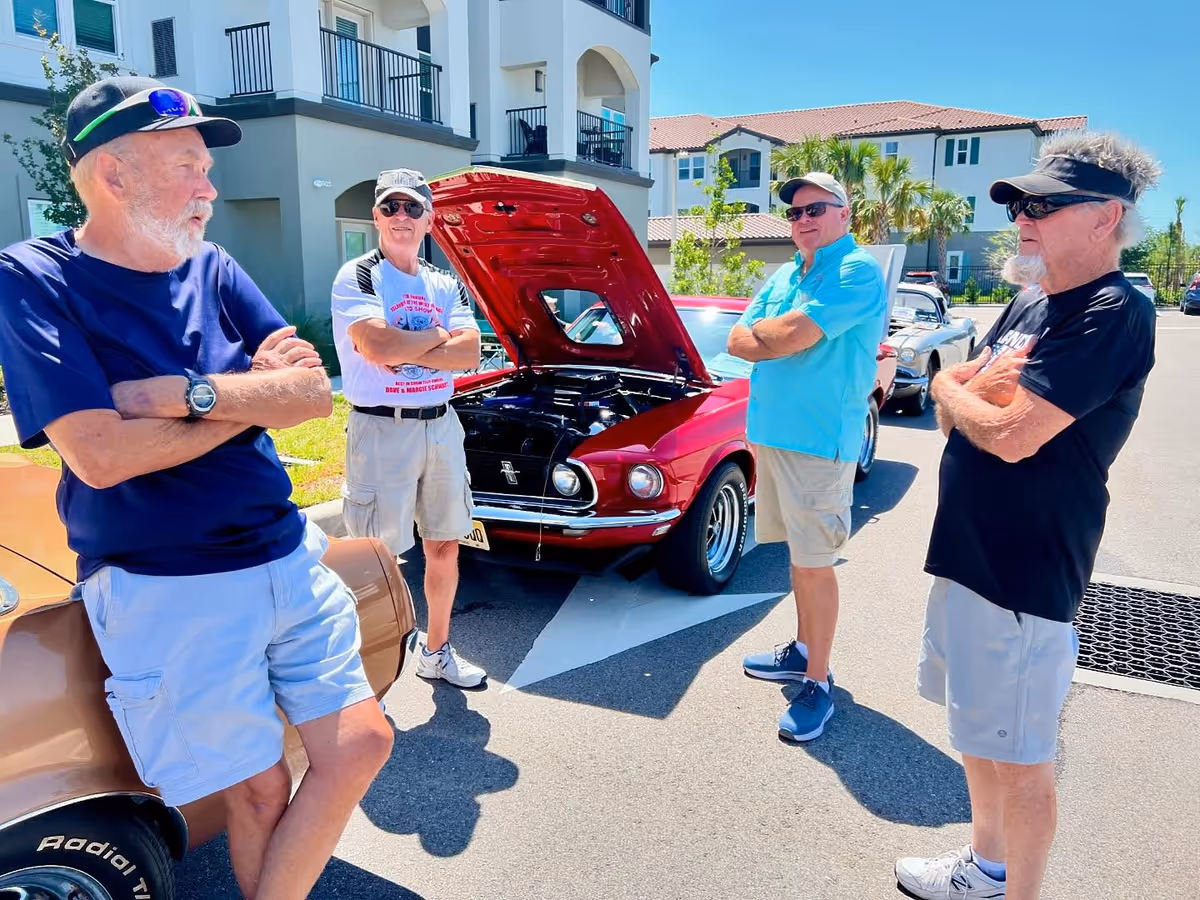Four older men standing outside near classic cars, with one red car's hood open. They are casually dressed in shorts and t-shirts, wearing sunglasses and hats, standing in a parking area in front of a residential building on a sunny day.