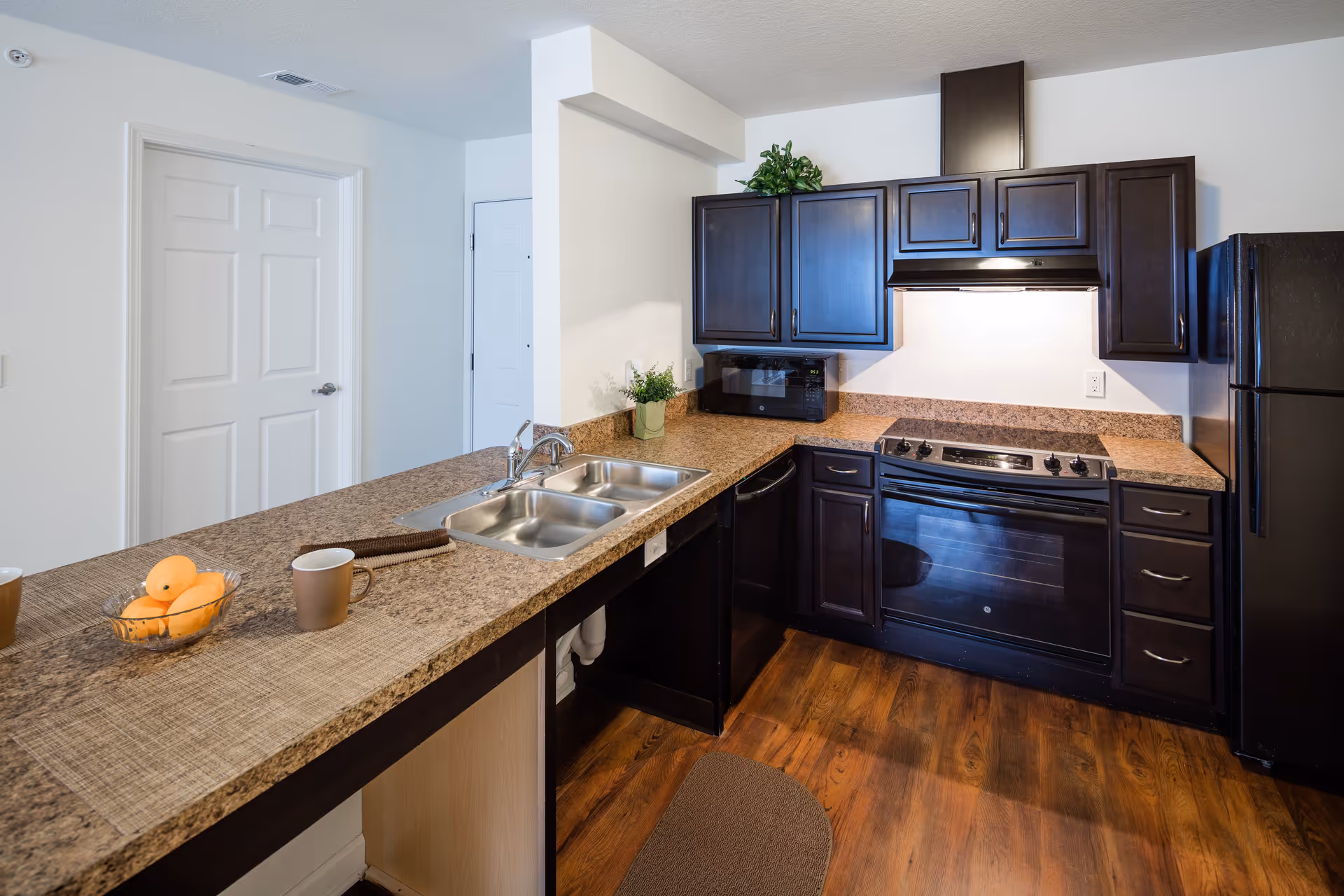 Modern kitchen with dark wood cabinets, a black refrigerator, black oven with stove, a microwave, and a double stainless steel sink set in a brown speckled countertop. The kitchen has wooden flooring and a small green plant on the counter. There are two brown mugs and a bowl of oranges on the counter with a beige placemat.
