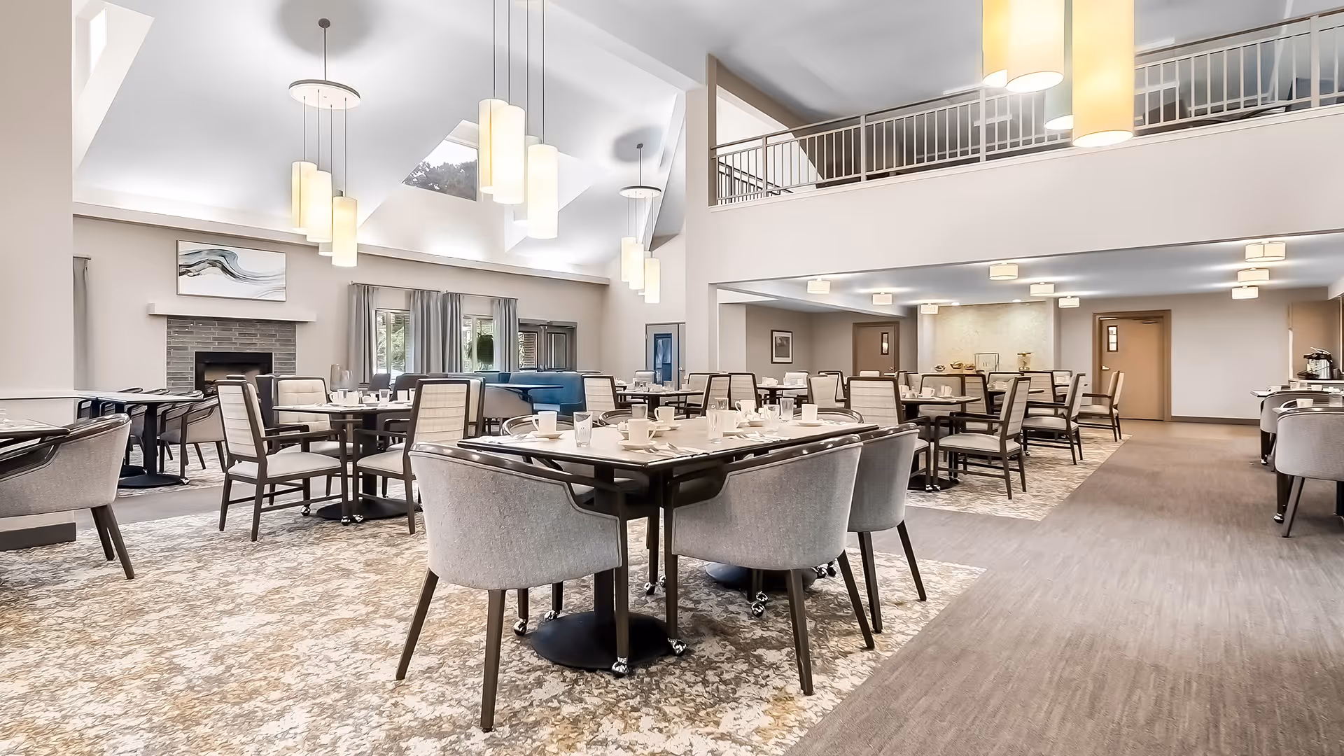 A spacious and well-lit dining room with multiple tables and chairs arranged neatly. The room features high ceilings with modern pendant lights hanging down, large windows with curtains, a fireplace with artwork above it, and a carpeted floor. The overall decor is contemporary and inviting.
