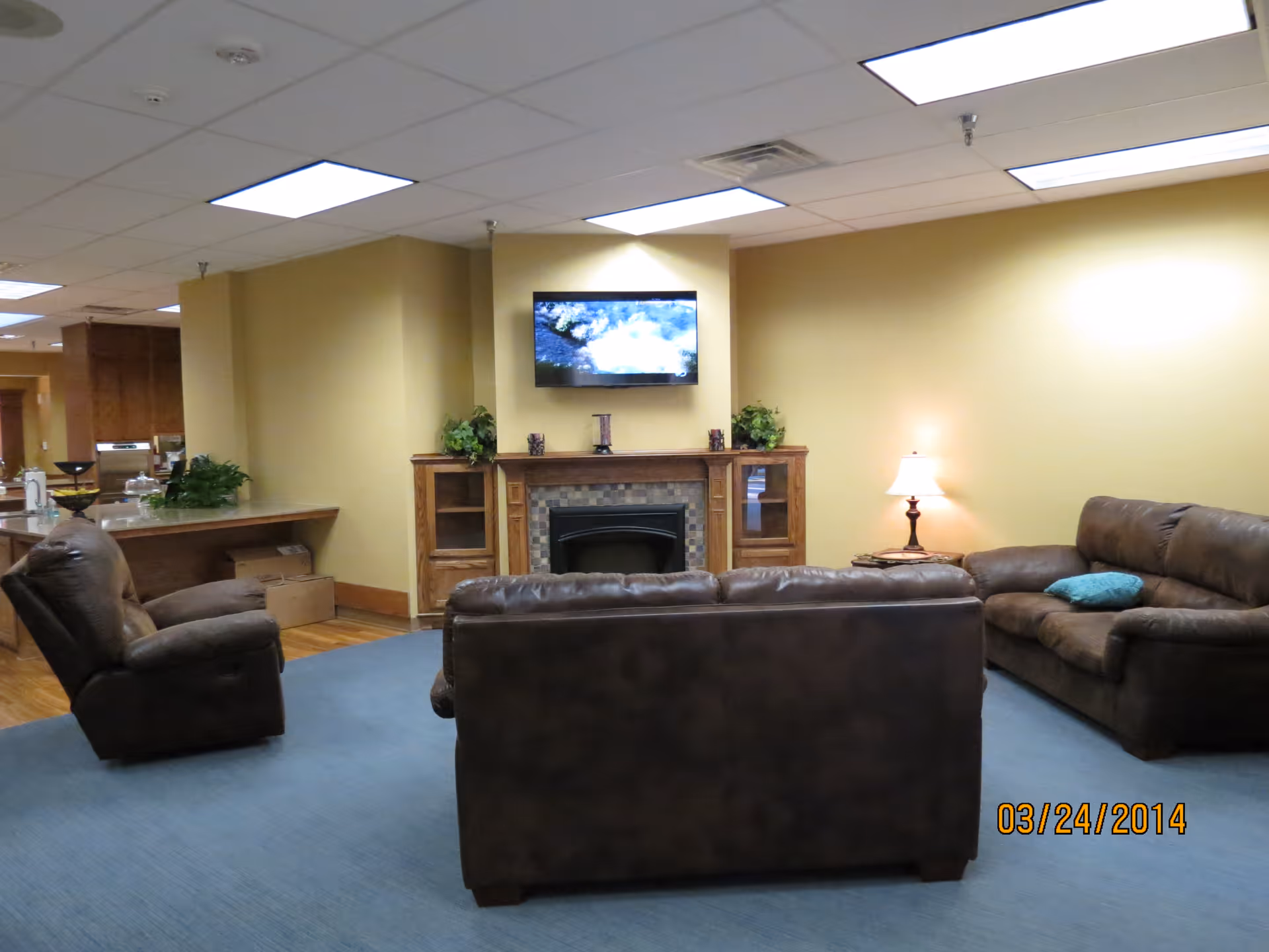 A cozy living room area with brown leather sofas and an armchair arranged around a fireplace with a mounted flat-screen TV above it. There are plants on the fireplace mantel and a table lamp on a side table next to one of the sofas. The walls are painted yellow and the floor is covered with a blue carpet. The ceiling has recessed lighting panels.