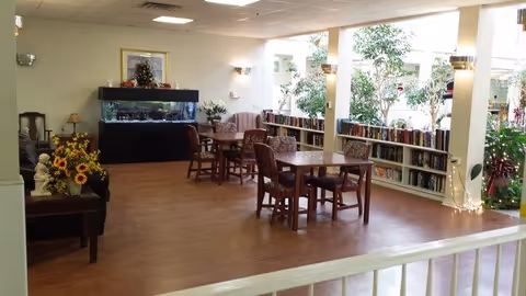 A bright and spacious common area with wooden floors, several tables and chairs arranged for seating, a large fish tank against the far wall, bookshelves filled with books along the right side, and large windows letting in natural light. There are plants and decorative lights around the room, creating a welcoming atmosphere.