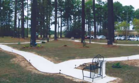 Winding white pathway through a grassy outdoor area with a bench and tall trees.