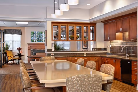 A modern kitchen and dining area with a large rectangular table surrounded by upholstered chairs. The kitchen features wooden cabinets, a granite countertop, a sink, and a dishwasher. Pendant lights hang above the table, and there is a view into a living room area with a fireplace and windows with blinds.