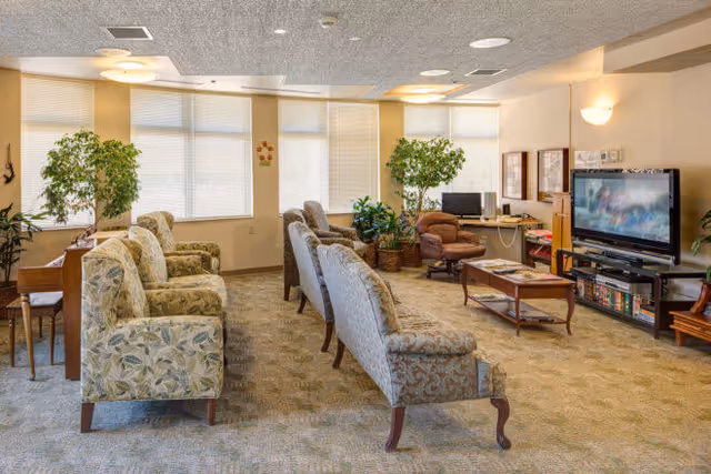 A spacious living room in an assisted living facility with multiple upholstered armchairs and sofas arranged facing a large flat-screen TV. The room has large windows with blinds, several potted plants, a coffee table with magazines, and a computer desk with a chair in the background.