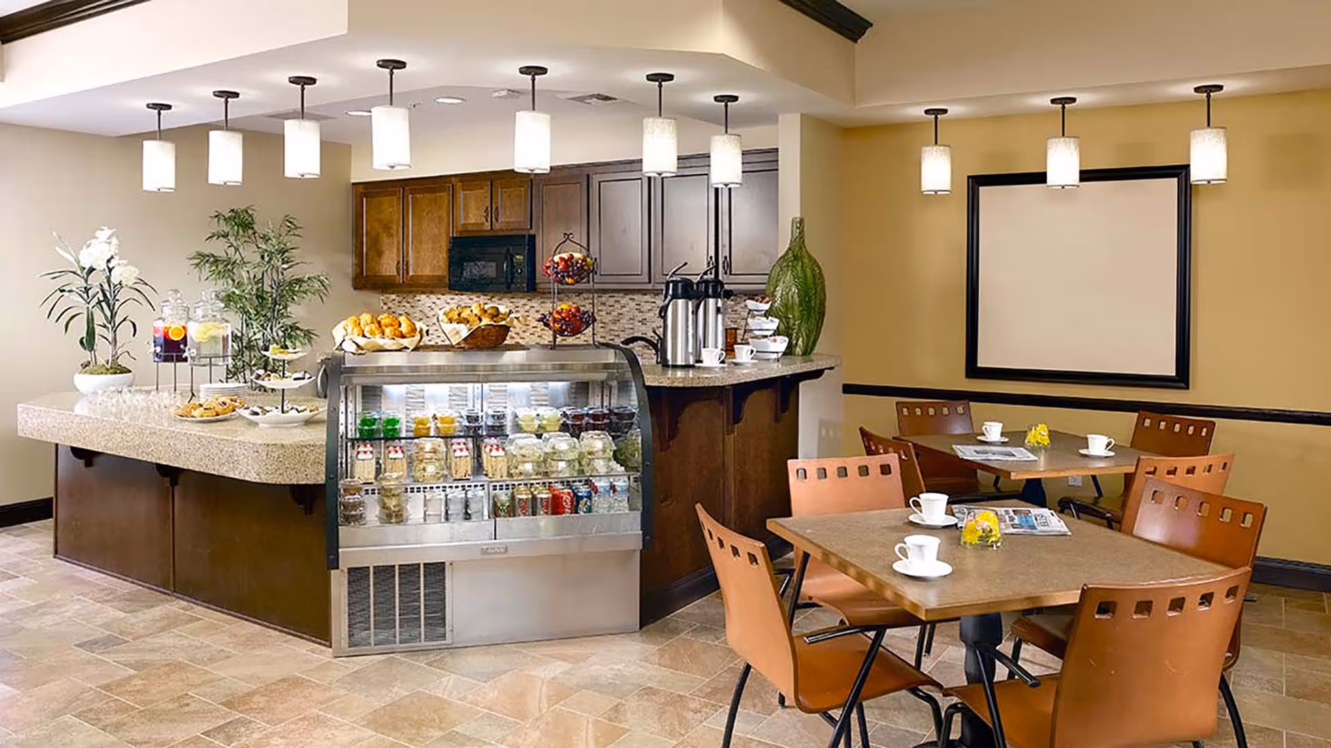 A cozy dining area with a small kitchen counter and refrigerated display case filled with beverages and snacks. The room features a beige and brown color scheme with pendant lights hanging from the ceiling. There are two tables with brown chairs, each set with white coffee cups and small yellow flower centerpieces. The kitchen counter has baskets of bread, coffee dispensers, and decorative plants.