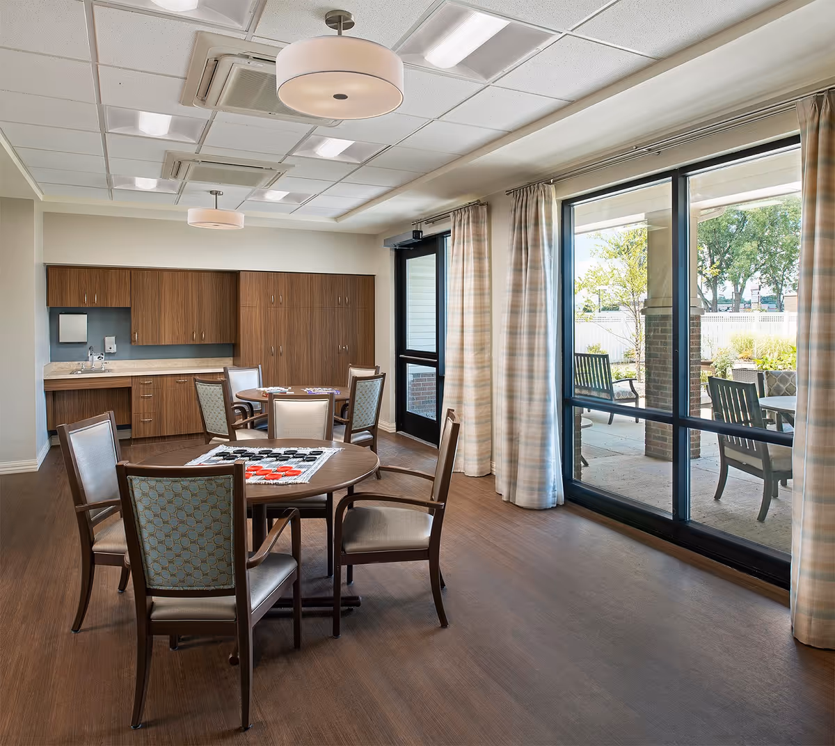 A bright room with wooden flooring featuring two round tables surrounded by chairs. One table has a checkers game set up. The room has large windows with curtains that look out onto a patio area with outdoor seating and greenery. There are wooden cabinets and a small sink area along one wall, and ceiling lights provide illumination.
