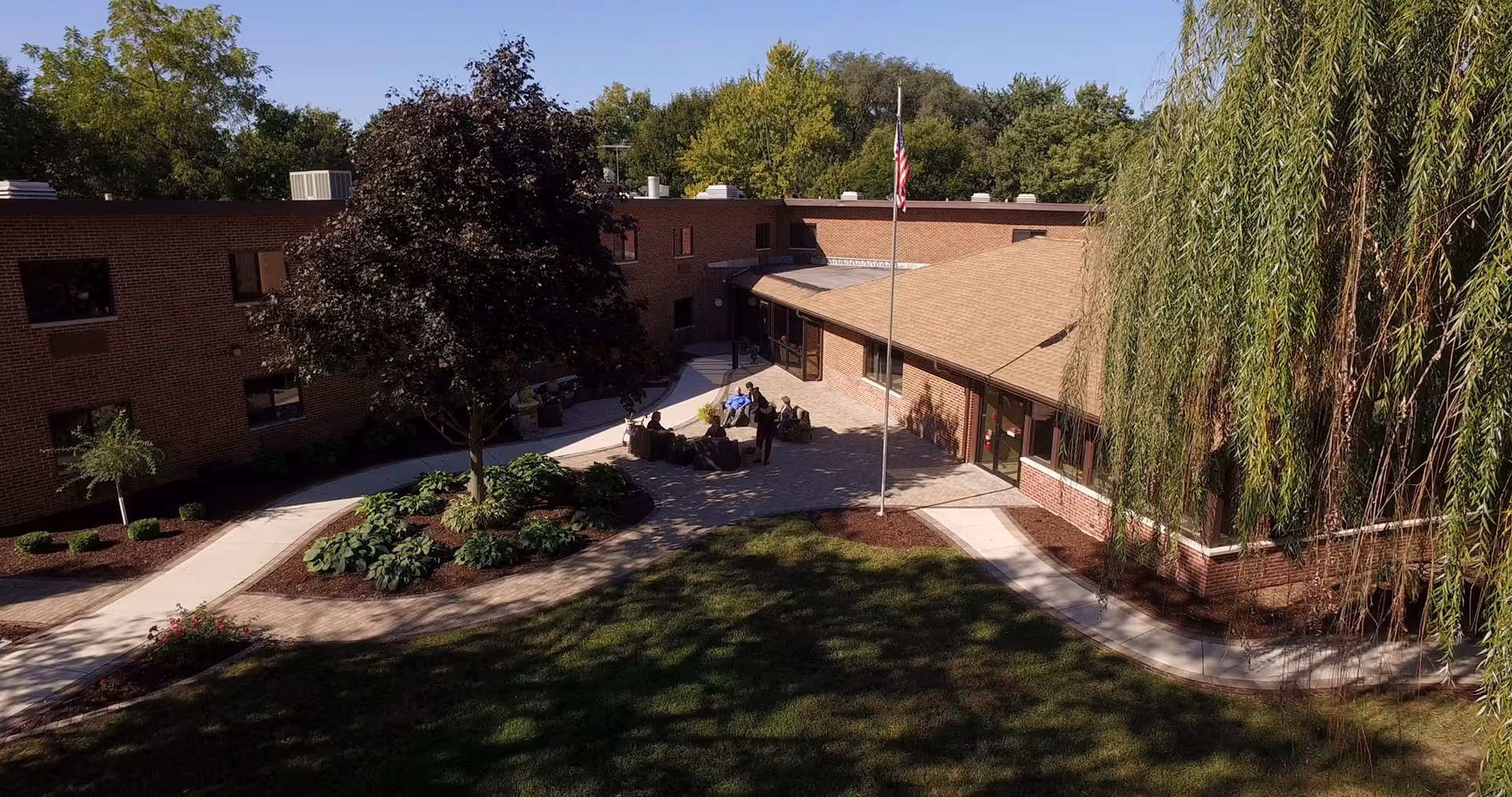Outdoor courtyard area of a senior living facility with a brick building surrounding it. There is a paved walkway, a flagpole with an American flag, several trees including a large weeping willow, and a group of people sitting and standing near outdoor furniture in the center of the courtyard.