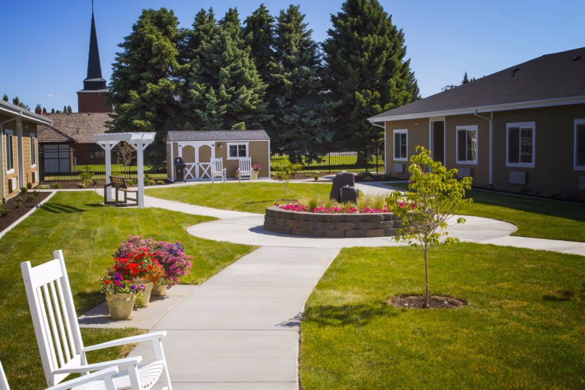 A well-maintained outdoor courtyard area with green grass, a circular flower bed with pink flowers, a small tree, white rocking chairs, potted flowers, a white pergola with a bench underneath, and a small shed in the background. The courtyard is surrounded by single-story buildings and tall evergreen trees, with a clear blue sky above.
