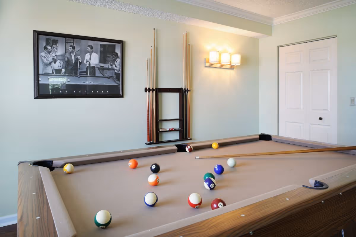 A pool table with billiard balls scattered on its tan felt surface in a room with light green walls. A rack holding pool cues is mounted on the wall above the table, next to a wall light fixture with three lamps. A black and white framed picture of The Rat Pack hangs on the wall. A white double door is visible in the background.